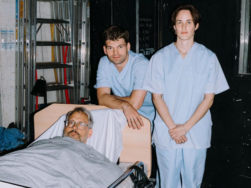 Martin Györffy und lynn t musiol tragen Krankenhauskittel und stehen nebeneinander. Christian Tschirner liegt neben ihnen in einem Krankenhausbett. 