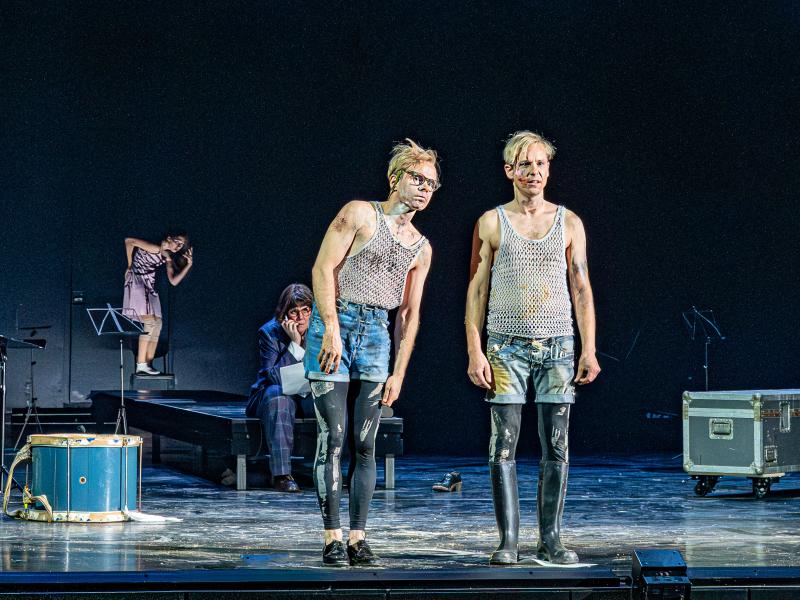 Kristof Van Boven und Nils Kahnwald stehen nebeneinander auf der Bühne. Kristofs Oberkörper neigt sich etwas in Richtung von Nils, die beiden blicken in dieselbe Richtung. Im Hintergrund sitzt Julia Wieninger auf einem Podest und beobachtet die beiden. Auf der Bühne verstreut stehen Notenständer, eine Kiste und eine Trommel. Weiter im Hintergrund tanzt eine Tänzerin.