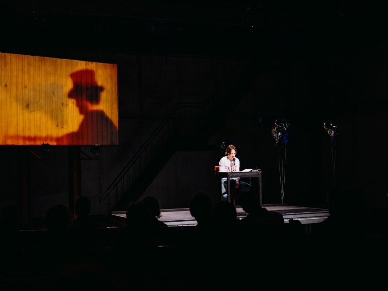 lynn t musiol sitzt auf der Bühne des MalerSaals an einem Tisch auf einem Stuhl, hält ein Mikrofon in der Hand und liest aus einem Buch. Im Hintergrund ist eine Leinwand zu sehen, auf der etwas verschwommen ein Mann mit Hut zu sehen ist. 