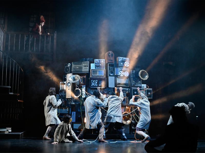 Aus mehreren Stereo- und Musikanlagen, Musikboxen und Grammophonen ist ein großer Gerätehaufen aufgebaut, vor dem fünf Personen stehen. Manche strecken die Arme nach dem Komplex aus, andere kämpfen mit Kabeln, die daraus kommen. Alberta von Poelnitz schaut von einem Balkon aus zu.