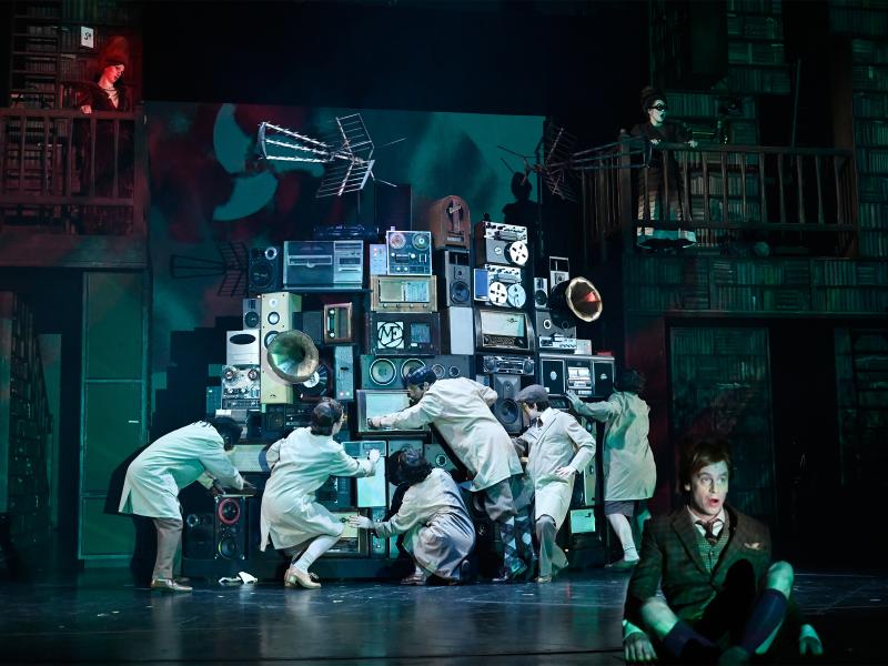 Aus mehreren Stereo- und Musikanlagen, Musikboxen und Grammophonen ist ein großer Gerätehaufen aufgebaut, vor dem fünf Personen stehen, die an den Anlagen rumdrehen. Jan-Peter Kampwirth sitzt im Schneidersitz mit geöffnetem Mund am Bühnenrand. Alberta von Poelnitz und Katja Danowski schauen von zwei Balkons im Hintergrund aus zu.