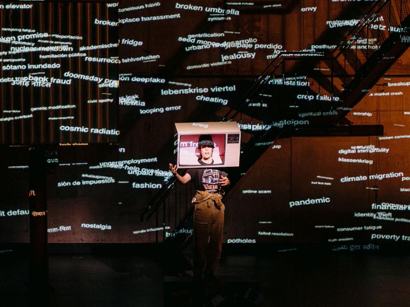 Wilson Tanner Smith steht auf der Bühne des MalerSaals. Auf seinem Kopf trägt er einen ausgeschnittenen Fernseher. Der Raum ist dunkel und wird nur von an die Wand projizierten Wörtern beleuchtet.  