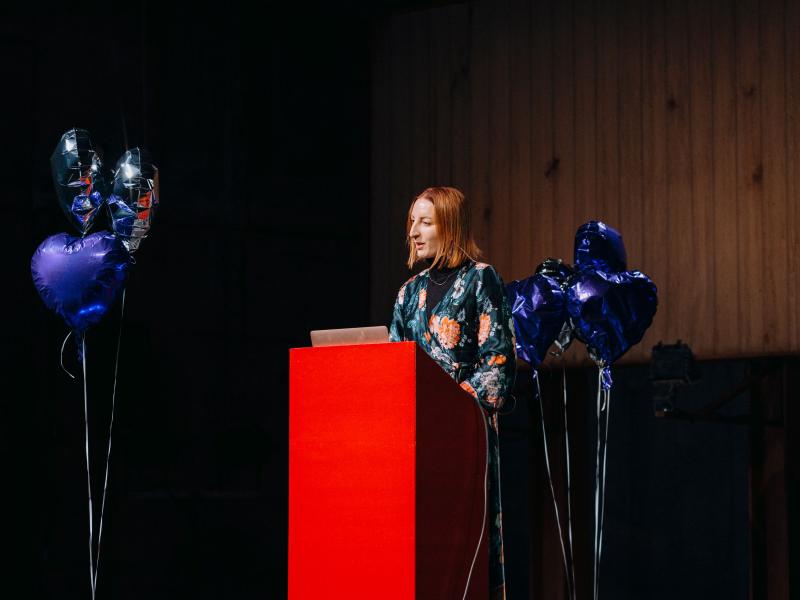 Dr. Ginie Servant-Miklos steht an einem roten Podium, um ihr herum sind silberne und blaue herzförmige Heliumluftballons.