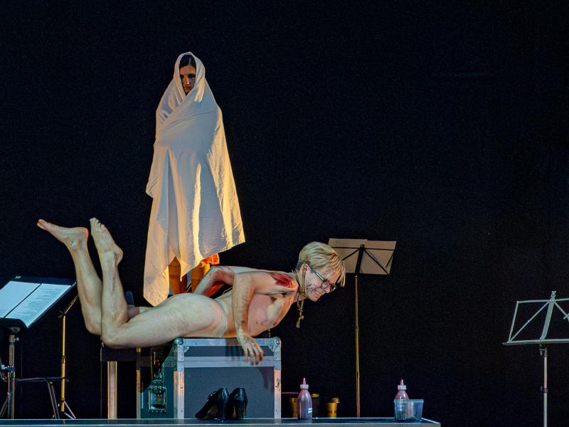 Kristof Van Boven liegt bäuchlings auf einer Kiste. Er ist nackt, seine Beine sind angewinkelt und er ist mit Blut beschmiert. Hinter ihm auf der Kiste steht eine Tänzerin in ein weißes Laken gehüllt, das nur ihr Gesicht zeigt und sie schaut Kristof Van Boven an. Um sie herum stehen Notenständer. Am Boden stehen ein Paar schwarze Absatzschuhe sowie Flaschen mit roter Flüssigkeit.