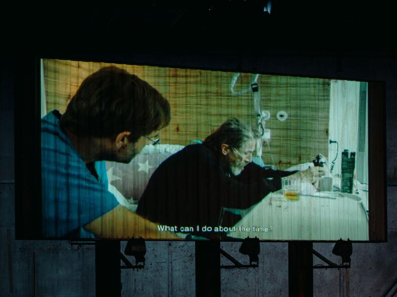 Auf der Bühne des MalerSaals steht eine Leinwand, auf der ein alter Mann in einem Krankenhausbett zu sehen ist. Neben ihm auf dem Bett sitzt ein anderer Mann im blauen Kittel. Als Untertitel steht dort „What can I do about the time?”