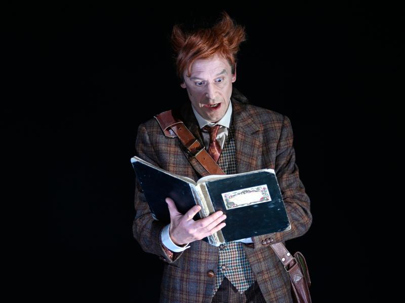 Jan-Peter Kampwirth steht alleine auf der dunklen Bühne. In den Händen hält er ein Buch, in das er mit geöffnetem Mund und weit aufgerissenen Augen schaut. Er trägt einen braun karierten Anzug mit kurzer Hose.