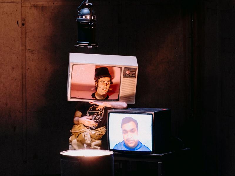 Wilson Tanner Smith steht auf der Bühne des MalerSaals. Auf seinem Kopf trägt er einen ausgeschnittenen Fernseher und lehnt auf einem echten Fernseher, auf dessen Bildschirm ein Mann zu sehen ist. 