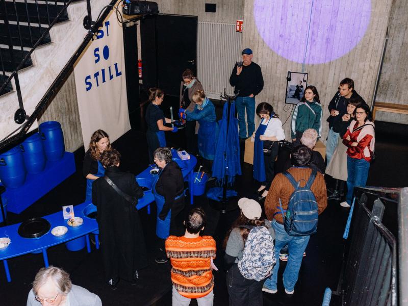 Viele Menschen stehen verteilt im MalerSaal. Auf einem blauen langen Tisch befinden sich Werkzeuge und Bastelutensilien. Manche Menschen tragen blaue Schürzen und Handschuhe.
