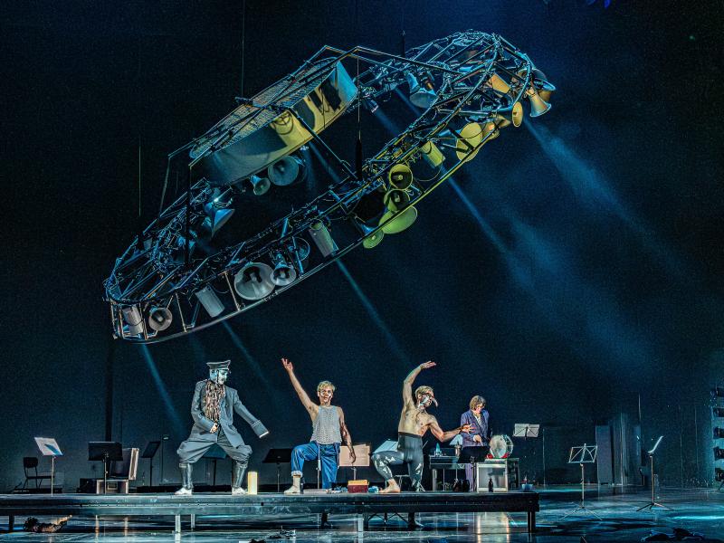 Drei Personen stehen nebeneinander hinter einem Podest und stellen einen Fuß darauf ab. Dabei strecken sie ihre Arme nach oben. Eine weitere Person steht breitbeinig auf dem Podest und trägt eine weiße Maske. Im Hintergrund stehen verteilt Notenständer auf der Bühne und eine Person neben einer Trommel. Über dem Bühnengeschehen hängt eine große kreisförmige Konstruktion aus verschiedengroßen und -artigen Lautsprechern.