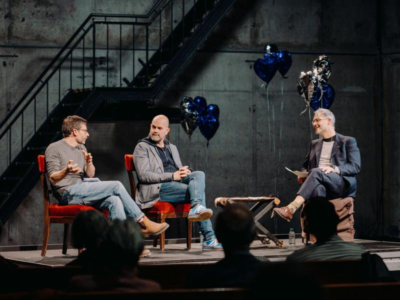 Christian Tschirner, Philipp Döring und Dr. Dirk Pörschmann sitzen nebeneinander in einem Halbkreis auf einem Podium. Sie gucken sich gegenseitig an. Im Hintergrund sind silberne und blaue herzförmige Heliumluftballons.