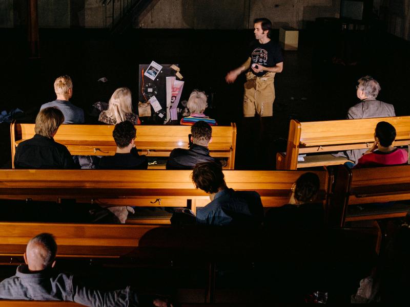 Wilson Tanner Smith steht mit einem Tischtennisball in der Hand auf der Bühne des MalerSaals. Er ist zum Publikum gewandt, das auf Kirchenbänken sitzt. 