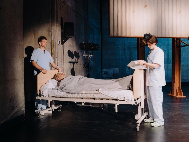 Martin Györffy und lynn t musiol tragen Krankenhauskittel und stehen um ein Krankenhausbett herum, in dem Christian Tschirner liegt. Sie befinden sich am Rand der MalerSaal Bühne. lynn t musiol durchblättert Zettel auf einem Klemmbrett.