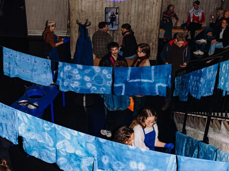 Viele Menschen stehen oder sitzen verteilt im MalerSaal. In der Mitte des Raumes steht ein blauer langer Tisch, auf dem sich Werkzeuge und Bastelutensilien befinden. An der Decke hängen Wäscheleinen, an denen blau gefärbte Tücher trocknen.  