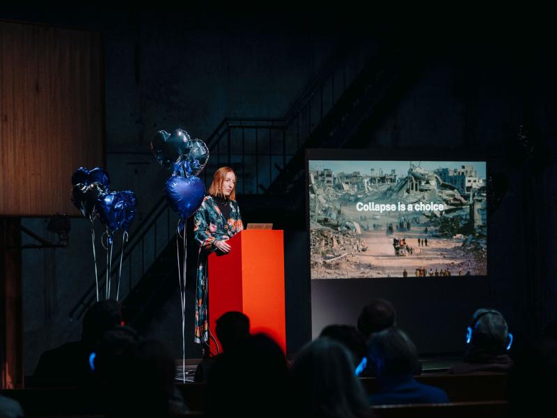 Dr. Ginie Servant-Miklos steht an einem roten Podium, um sie herum sind silberne und blaue herzförmige Heliumluftballons. Neben ihr wird ein Kriegsbild mit der Aufschrift „Collapse is a choice“ auf eine Leinwand projiziert.