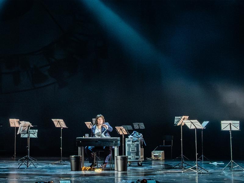 Julia Wieninger sitzt in einem lila-grün-karierten Anzug mit weißem Hemd an einem Tisch. Neben dem Tisch stehen zwei schwarze Mülleimer. Ihre linke Hand ist erhoben, ihr Mund ist geöffnet. Um sie herum stehen viele Notenständer quer im Raum verteilt.