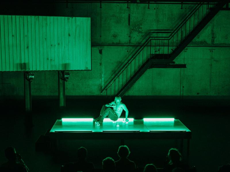 Auf einem grün beleuchteten Podium auf der Bühne des MalerSaals balanciert ein Mann mit den Beinen, dem Po und einem Arm auf umgedrehten Weingläsern. 