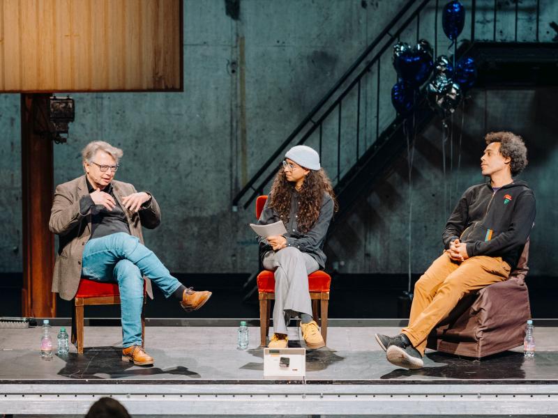 Prof. Dr. Jürgen Zimmerer, Kira Herff und Amewu sitzen nebeneinander in einem Halbkreis auf einem Podium auf Stühlen. Sie gucken sich gegenseitig an. Um sie herum sind silberne und blaue herzförmige Heliumluftballons. 
