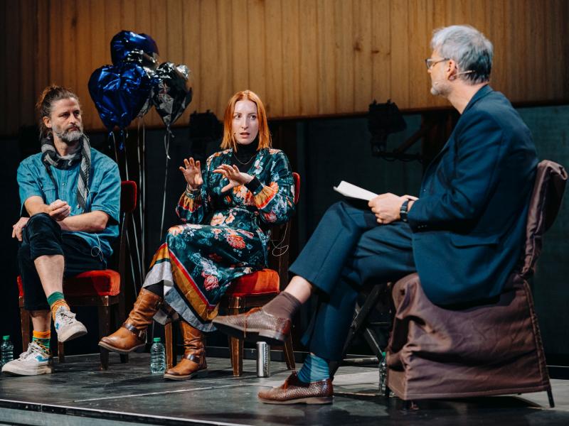 Christophe Meierhans, Dr. Ginie Servant-Miklos und Christian Tschirner sitzen nebeneinander in einem Halbkreis auf einem Podium. Dr. Ginie Servant-Miklos gestikuliert mit ihren Händen, die Männer schauen sie an. Im Hintergrund sind silberne und blaue herzförmige Heliumluftballons zu sehen.