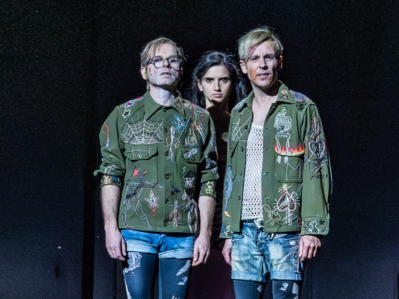 Kristof Van Boven und Nils Kahnwald stehen nebeneinander auf der Bühne. Sie tragen schwarze Leggings mit weißen Mustern, kurze Jeanshosen darüber und dunkelgrüne Jacken mit Symbolen und Bildern, wie einem Spinnennetz, Sternen, Feuer, Herzen, Skeletten. Hinter ihnen mittig steht Maria Carolina Vieira und schaut gerade zwischen ihnen hindurch.