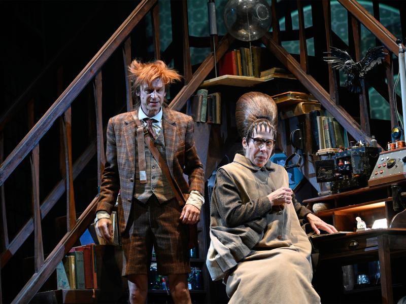 Jan-Peter Kampwirth steht neben Ute Hannig, die an einem Schreibtisch sitzt. Beide sind von Holzregalen mit Büchern umgeben.