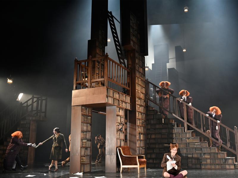 Drei Personen in Daunenjacken-Kleidern und mit orangen aufgeplusterten Perücken stehen nebeneinander auf einer Treppe eines großen Bücherregals. Im Hintergrund sind Tänzer*innen zu sehen. Jan-Peter Kampwirth sitzt im Schneidersitz mit geöffnetem Mund am Bühnenrand und schreibt etwas mit einer Feder auf ein Blatt Papier. 