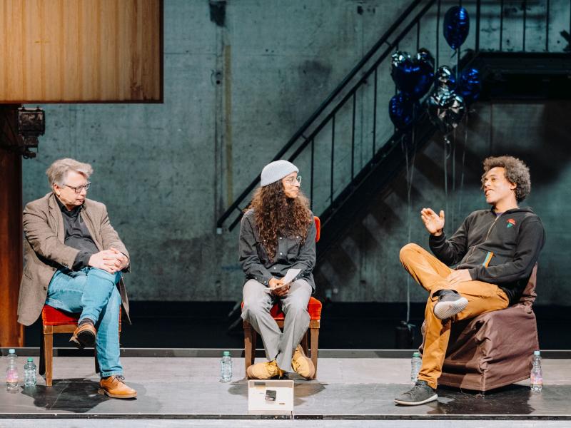 Prof. Dr. Jürgen Zimmerer, Kira Herff und Amewu sitzen nebeneinander in einem Halbkreis auf einem Podium auf Stühlen. Sie gucken sich gegenseitig an. Um sie herum sind silberne und blaue herzförmige Heliumluftballons. 