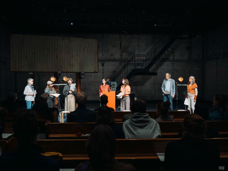 Diverse Performer*innen stehen gemeinsam auf der MalerSaal Bühne. Sie sprechen miteinander und hören sich gegenseitig zu. Das Publikum ist von hinten zu sehen. 