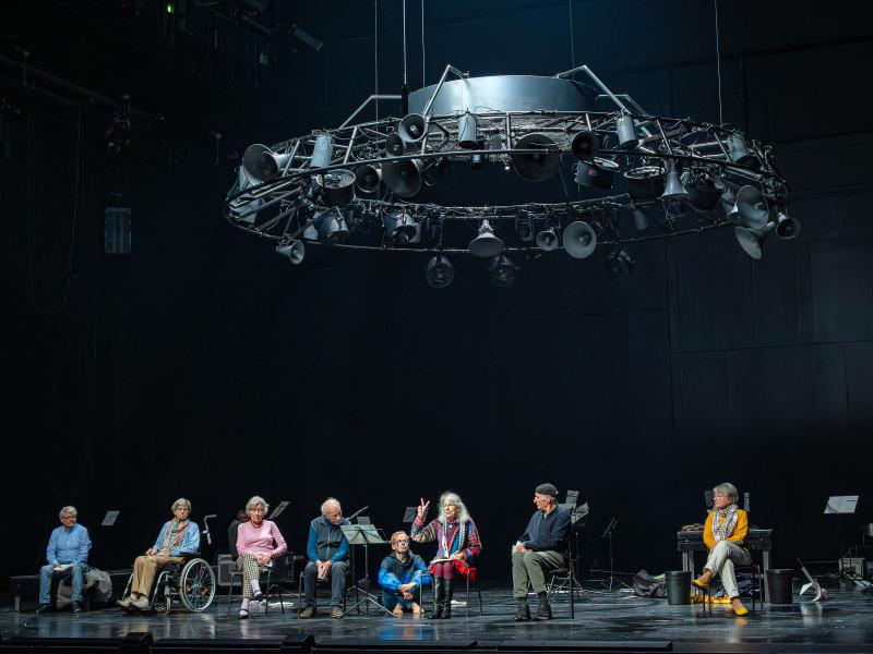 Alle sieben Zeitzeug*innen der Inszenierung sitzen nebeneinander auf der Bühne. Eine Person mittig hat eine Hand gehoben und Mittel- und Zeigefinger gespreizt. Alle schauen zu ihr. Rechts steht Julia Wieninger an einem Mikrofon, Kristof Van Boven sitzt zu den Füßen der sprechenden Person und schaut sie an. Über der Szene hängt eine große runde Stahlkonstruktion aus vielen unterschiedlich großen alten trichterförmigen Lautsprechern, Scheinwerfern und Ventilatoren.