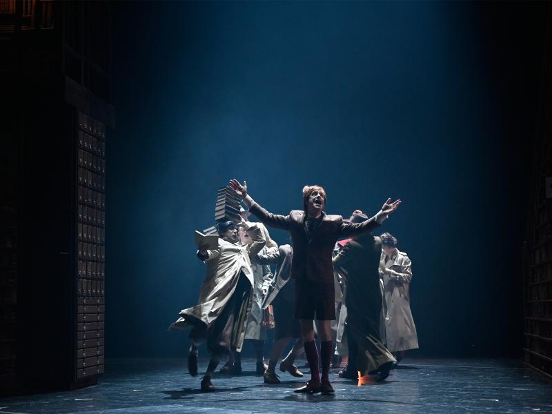 Jan-Peter Kampwirth steht mit ausgestreckten Armen und geöffnetem Mund im Vordergrund, hinter ihm sind mehrere Personen in Bewegung, die Trenchcoats tragen. Einer Frau fallen gerade Bücher aus der Hand.