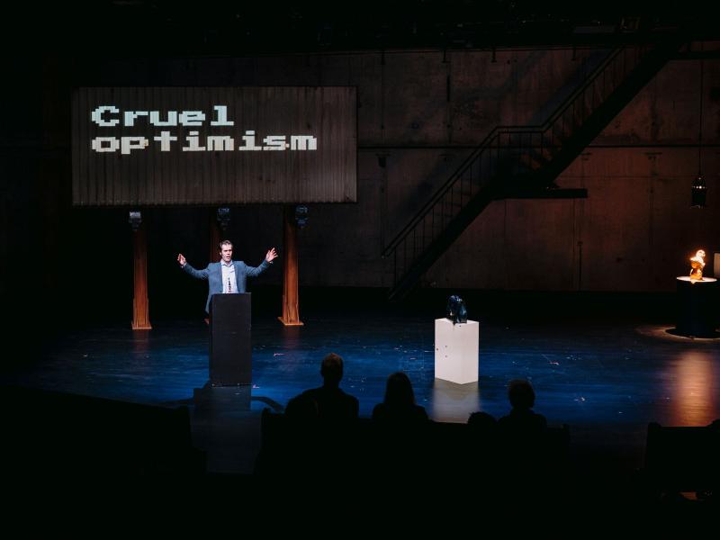 Wilson Tanner Smith steht auf der Bühne des MalerSaals an einem Podium. Er streckt die Arme zu den Seiten aus. Im Hintergrund ist eine Leinwand zu sehen, mit der Aufschrift „Cruel optimism“. 