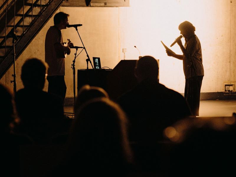 Zwei Performer*innen stehen sich auf der MalerSaal Bühne gegenüber. Die rechte Person hält ein Mikrofon und einen Zettel in der Hand, die linke Person spielt Ukulele. Die Lichtverhältnisse lassen nur zu, dass man die Schatten der Performer*innen sieht. Das Publikum ist von hinten zu sehen.