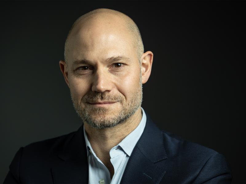 Omri Boehm steht vor einem dunklen, neutralen Hintergrund. Er trägt einen dunklen Anzug und lächelt in die Kamera.