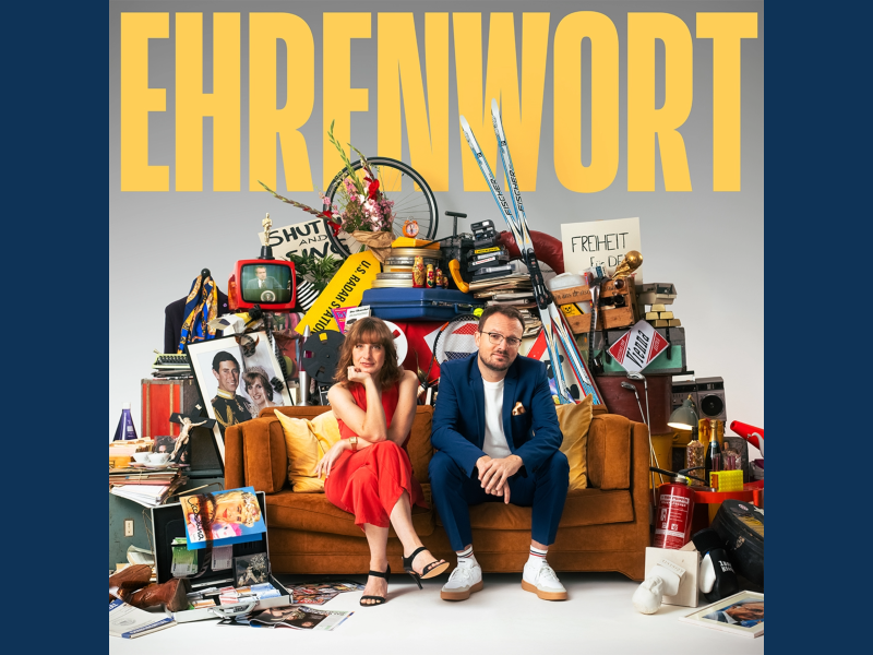 Das Cover des Ehrenwort-Podcasts. Oben drüber steht in großen gelben Buchstaben "EHRENWORT". Auf einem orangenen Sofa sitzen eine Frau in einem roten Kleid und ein Mann mit einem blauen Anzug an. Sie sind umgeben von zahlreichen Gegenständen, wie zum Beispiel Skiern, einem Fahrradreifen, einem Bilderahmen, einem Feuerlöscher und vielen weiteren Dingen.