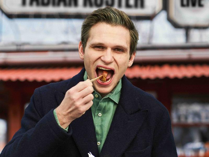 Fabian Köster träg ein grünes Hemd und eine blaue Jacke. Er führt eine Hand mit einem kleinen Spieß zum Mund, an dem ein Stück Currywurst aufgespießt ist.