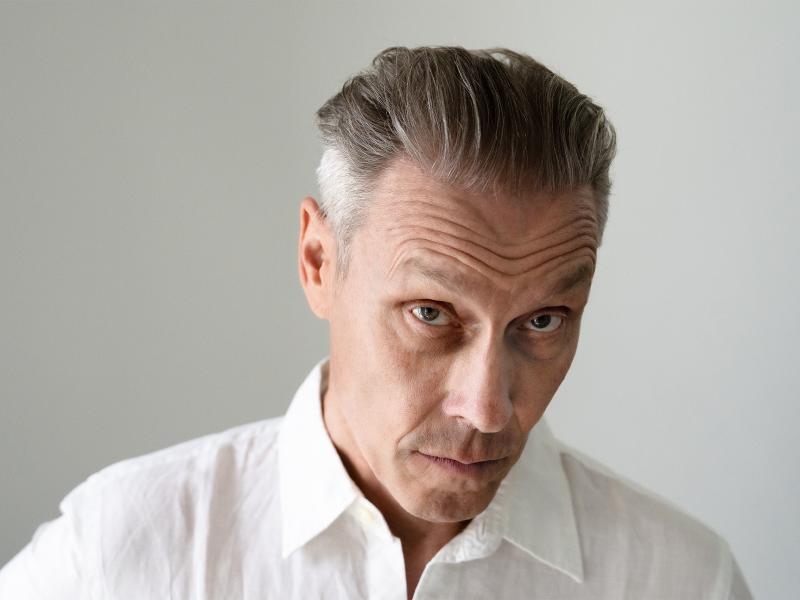 Farin Urlaub ist vor einem neutralen Hintergrund zu sehen. Er trägt ein weißes Hemd und kurze graue Haare. Er schaut in die Kamera.