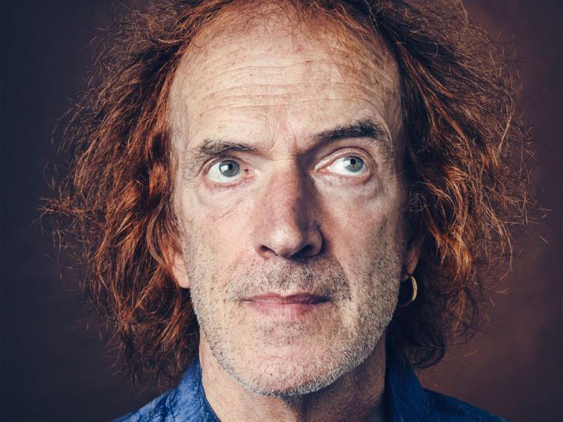 Graham Valentine steht vor einem neutralen Hintergrund. Er trägt ein hellblaues Hemd und kinnlange rote Haare. Er schaut nach oben, an der Kamera vorbei.