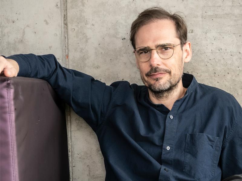 Jochen Schliemann sitzt auf einer lilafarbenen Couch vor einem neutralen Hintergrund. Er trägt ein dunkelblaues Hemd, eine dunkle Brille und kurze, braune Haare. Er schaut in die Kamera.