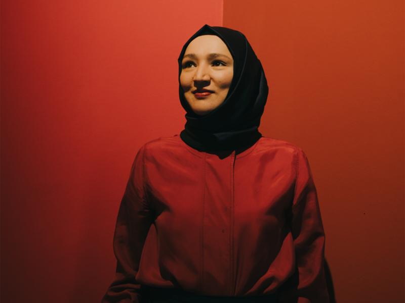 Kübra Gümüşay steht vor einem roten Hintergund. Die trägt ein rotes Oberteil und ein schwarzes Kopftuch. Sie lächelt und schaut leicht an der Kamera vorbei.