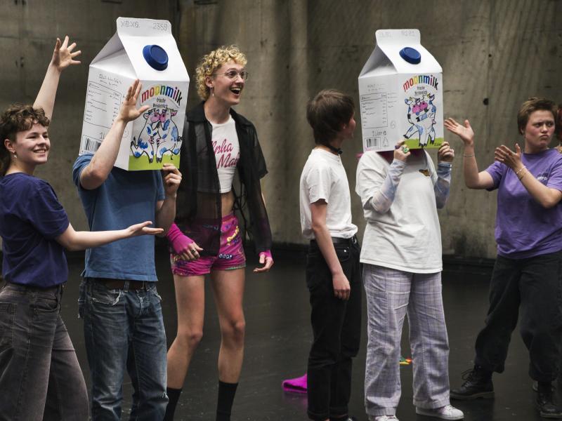 Sechs Jugendliche stehen nebeneinander in einer Reihe. Zwei von ihnen tragen Milchpackungen auf dem Kopf, sodass man ihre Gesichter nicht mehr erkennt. Auf den Milchpackungen sind jeweils zwei Kühe zu erkennen, die sich küssen. Die Personen, deren Gesichter man erkennen kann, lächeln und strecken ihre Arme in Richtung der Personen, die verkleidet sind.