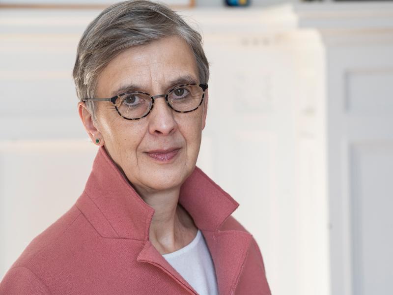Katharina Pistor steht vor einem neutralen Hintergrund. Sie trägt kurze, graue Haare, eine dunkle Brille und ein rosafarbenes Oberteil. Sie schaut in die Kamera.