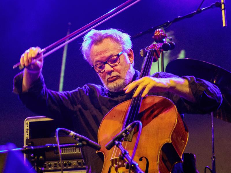 Konzertsituation. Martin Schütz trägt ein schwarzes Oberteil, eine dunkle Brille und kurze graue Haare. Im Hintergrund ein Konzertaufbau und blaues Bühnenlicht. Er hält ein Cello und hebt den Bogen zum Spielen.