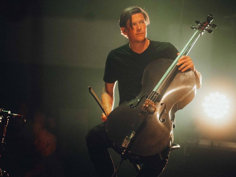 Konzertsituation: Niklas sitzt in einem schwarzen T-Shirt auf einem Stuhl und hält ein Cello in der Hand. Er schaut leicht nach oben und hat den Bogen in der Hand. Von hinten scheint Gegen licht in die Kamera.