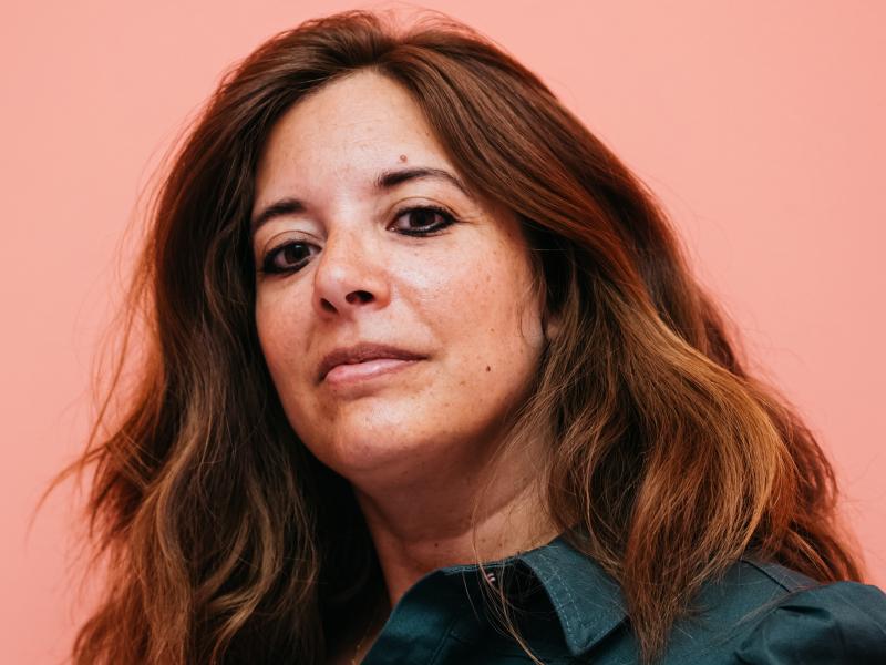 Rasha Khayat steht vor einem neutralen Hintergrund. Sie trägt offene, schulterlange Haare und ein dunkelgrünes Hemd. Sie schaut in die Kamera.