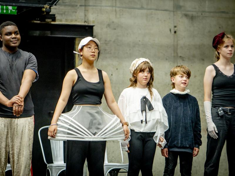 Fünf junge Schauspieler*innen stehen nebeneiner in einer Reihe auf der MalerSaal Bühne. Zwei von ihnen sind deutlich kleiner und jünger als die drei anderen. Eine trägt einen ausgefallenen durchsichtigen weißen Rock, die anderen sind dunkel gekleidet.