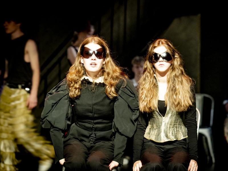 Zwei junge Schauspielerinnen sitzen nebeneinander auf der MalerSaal Bühne. Sie tragen dunkle Kleidung und sehr große Sonnenbrillen. Im Hintergrund sind noch weitere Schauspieler*innen zu sehen, die sich bewegen.