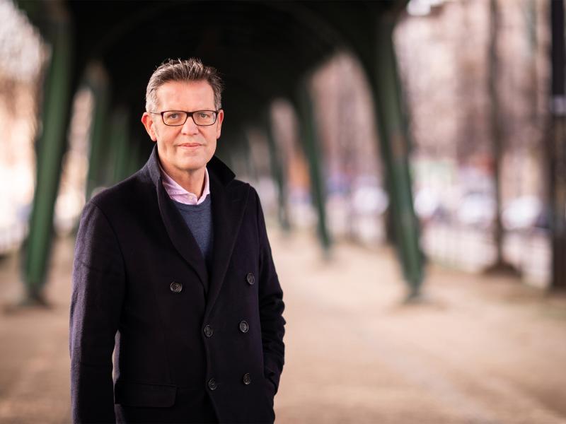 Steffan Mau steht draußen unter einer Brücke, die aussieht, wie eine Hochbahnbrücke. Er trägt eine dunkle Jacke, kurze graue Haare und eine Brille. Er schaut in die Kamera.