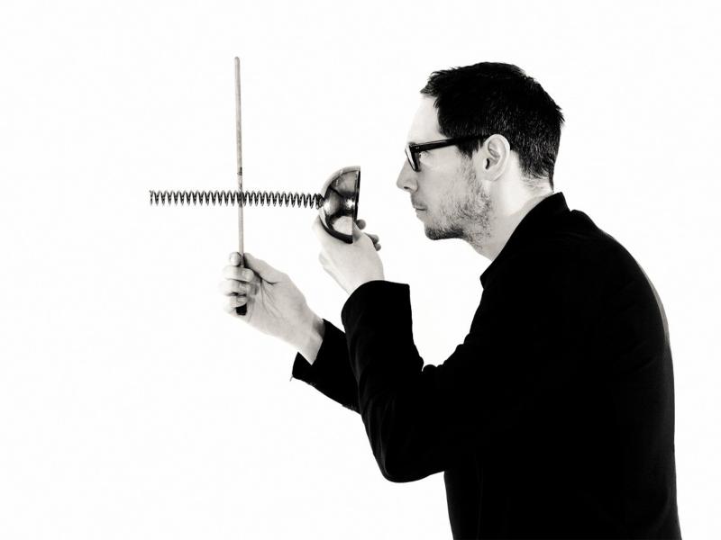 Schwarz-Weiß_Foto. Stephan Krause-Bantzer steht vor einem weißen Hintergrund im Profil. Er trägt ein schwarzes Hemd und eine dunkle Brille. Er hält zwei metallene lange Federn in beiden Händen und hält sie vor sein Gesicht.