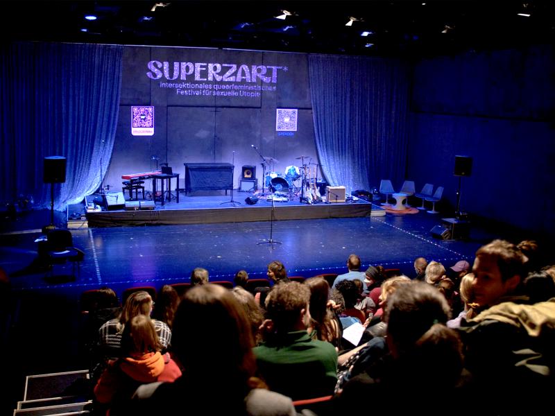 Eine Bühne ist mit Musikinstrumenten eingerichtet. Rechts und links von der Bühne hängt ein heller Vorhang. Über der Bühne ist der Schriftzug "SUPERZART* - intersektionales queerfeministisches Festival für sexuelle Utopie" zu lesen. Vor der Bühne sitzen viele Menschen auf roten Theatersitzen.