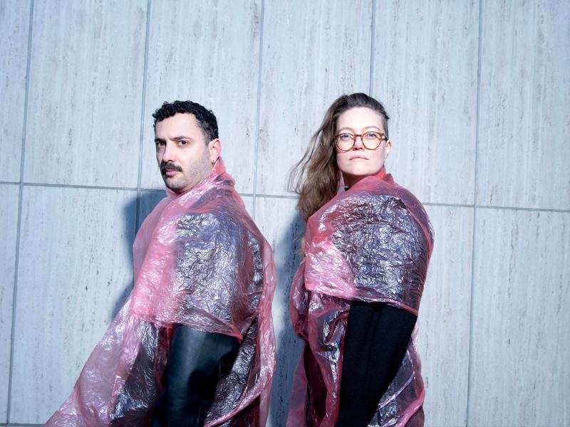 Fikri Anıl Altıntaş und Katharina Wisotzki stehen vor einer grauen Wand. Beide schauen in die Kamera und tragen schwarze Plastikanzüge mit rosafarbenen Regencapes drüber.