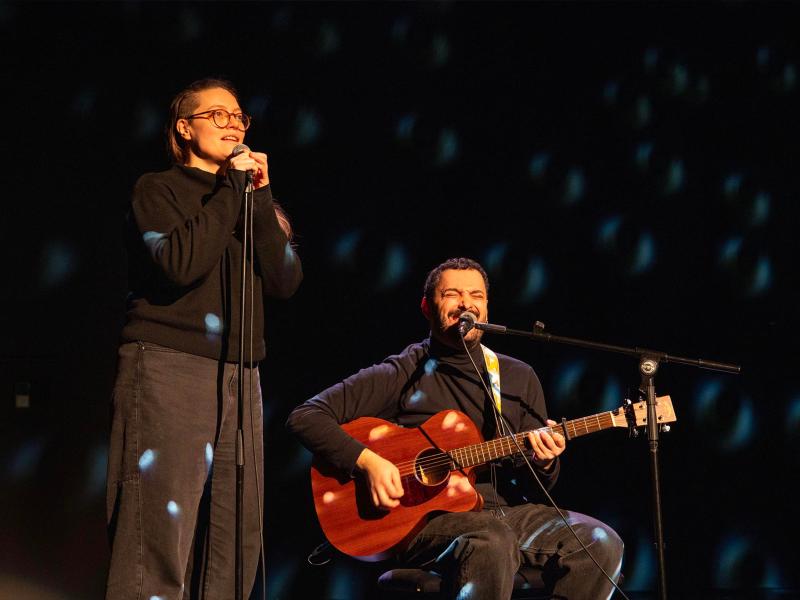 Bühnensituation. Fikri Anıl Altıntaş sitzt auf einem Stuhl, spielt auf einer roten Gitarre und sind in ein stehendes Mikrofon. Katharina Wisotzki steht daneben und spricht oder singt ebenfalls in ein Mikrofon. Beide tragen schwarze Kleidung.